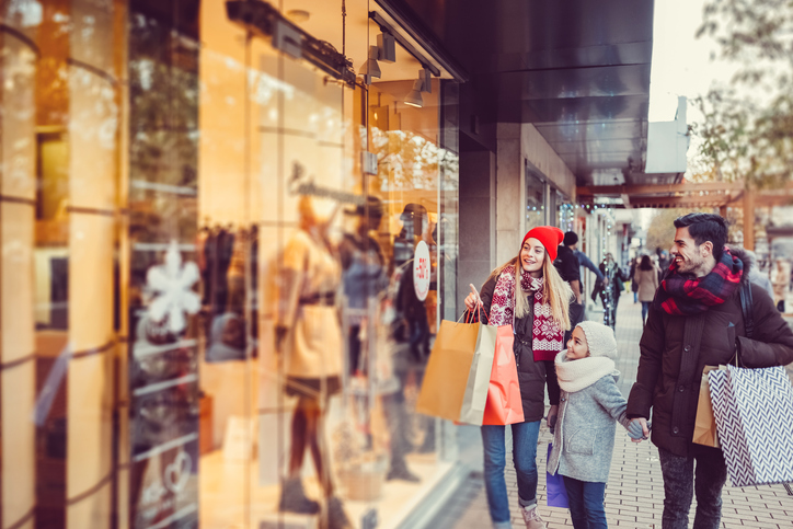 Do lado direito da imagem tem um homem, uma mulher e uma criança segurando algumas sacolas de compras e olhando para uma vitrine de roupas que está localizada do lado esquerdo da imagem.