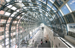 A imagem mostra o interior do Eaton Centre Toronto contemplando três andares do shopping. À esquerda, podem ser vistas as engrenagens do elevador central e, à direita, alguns transeuntes observando as lojas. No centro da imagem, um corredor pode ser visto abarrotado de gente. Acima, se vê o famoso teto de vidro do complexo de lojas.