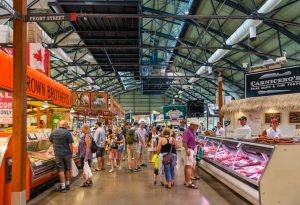 A imagem é do St. Lawrence Market, o mercado público de Toronto. Nela é possível ver pessoas andando e olhando para os stands de comidas no interior do mercado.