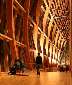 A imagem mostra um dos corredores da parte interior da AGO. O piso de madeira e as colunas possuem a mesma coloração ocre. Algumas pessoas estão na imagem, caminhando e sentadas em bancos distribuídos no corredor