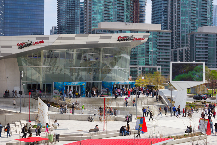 Essa é a fachada do Ripley’s Aquarium of Canada, aquário de Toronto. No Fundo da imagem tem alguns prédios espelhados e a frente está prédio do aquário de Toronto e uma escadaria na frente com pessoas espalhadas, algumas na porta do local e outras descendo a escadaria.