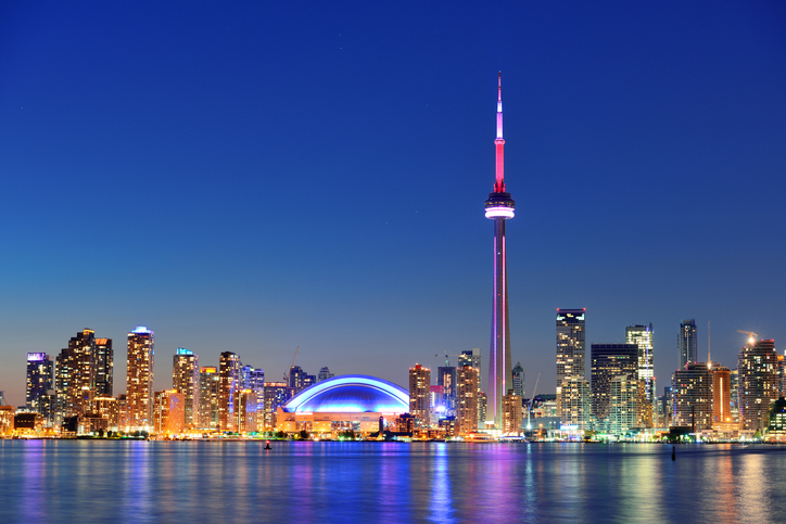 A imagem contempla uma visão panorâmica da cidade de Toronto. Na parte da frente é possível ver o lado Ontário e ao longe prédios iluminados em uma paisagem noturna. A CN Tower, maior torre de Toronto, está localizada próxima ao lado direito da imagem com uma iluminação rosada.