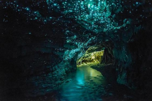 A imagem mostra a caverna Waitomo. Um leito de rio subterrâneo corre para o fundo da imagem, ao centro. Grande parte do teto e algumas partes da parede são tomadas por milhares de vagalumes azuis que dão luminosidade à caverna, produzindo um bonito efeito visual.