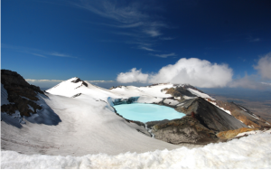 Descrição da imagem: Em um dos cumes da montanha Tongariro, fica o lago de mesmo nome. Dependendo da temperatura, os microrganismos que vivem no lago proporcionam um espetáculo de cores que pode ser azul bem claro (como na imagem descrita) ou em tons de verde esmeralda. O lago é cercado por montes de neve, e o céu está bem claro.