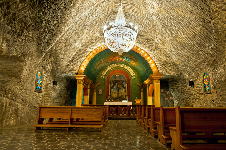  Descrição da imagem: A imagem mostra uma das capelas presentes nas minas de sal de Wieliczka, com o teto, paredes e piso igualmente acinzentados por conta do sal. Um grande lustre ilumina as imagens cristãs do ambiente, juntamente com os bancos de madeira.