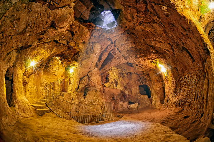 Descrição da imagem: na imagem, a cidade subterrânea Turquia, na Capadócia. Um túnel feito de pedra vulcânica com diversas entradas e saídas nas laterais da imagem. Uma fenda no teto, no centro da imagem, permite a entrada de luz natural em Derinkuyu.