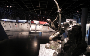 A imagem mostra a parte de dentro do Museu Olímpico Lausanne, na Suíça. Há uma escultura de um homem agachado com um arco que aponta para o alto com a sua flecha. Ao fundo, réplicas de tochas olímpicas históricas decoram uma das seções do museu suíço.
