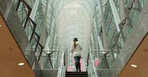 Descrição da imagem: podemos observar na imagem uma das saídas do sistema PATH cidade subterrânea Toronto, onde uma escada rolante central leva uma mulher para um dos túneis acima da terra. O túnel possui o teto tomado por vidros, o que proporciona uma belíssima iluminação natural.