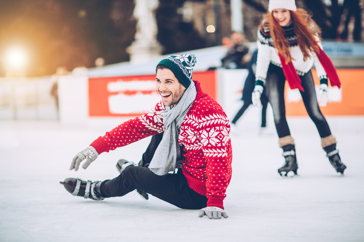 Na imagem, há um homem e uma mulher em uma pista de patinação de gelo. Os dois estão vestidos para o inverno com gorro, cachecol, calça, blusa de lã (ele de vermelho e ela de branco e preto) e patins para patinação no gelo. O homem está sentado no chão congelado e a mulher está indo em direção a ele. Os dois estão sorrindo.