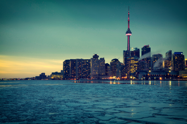 A fotografia contempla o lago Ontário congelado e, ao fundo, os prédios da cidade de Toronto iluminados em um começo de noite na região. A CN Tower, maior torre do Canadá, está no meio desses mesmos prédios com uma iluminação rosada.
