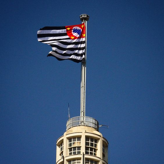 escrição da imagem: a imagem mostra a bandeira do estado de São Paulo tremulando no alto do edifício Altino Arantes, popularmente conhecido como Banespão.