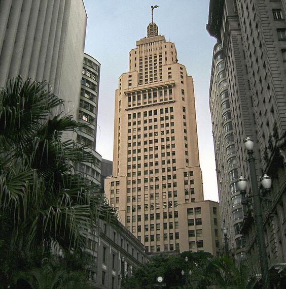 a fotografia foi tirada de baixo, mostrando o imponente Banespão em meio aos prédios do centro velho da cidade de São Paulo. A bandeira do estado tremula de cima dos seus 35 andares.