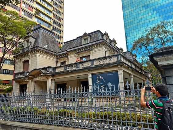 Descrição da imagem: a imagem apresenta a Casa das Rosas sendo vista da Avenida Paulista. O casarão de dois andares está atrás de portões de ferro. Um casal é fotografado em uma das sacadas do piso superior.