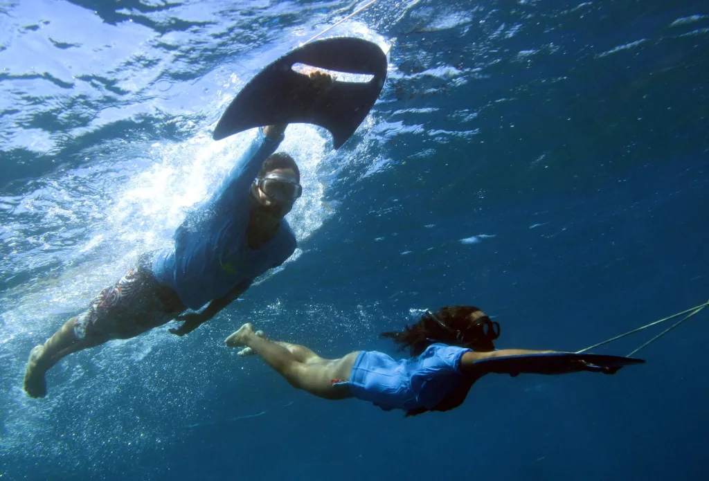 Descrição da imagem: Duas pessoas se aventuram no Aquasub, segurando a pequena prancha na mão e utilizando máscaras e snorkel.