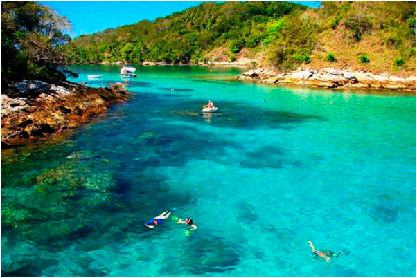 A foto mostra os entornos da Ilha Grande em Angra dos Reis. Um mar de água cristalina convida banhistas a nadarem entre as pedras. Um barco ao fundo espera os turistas.