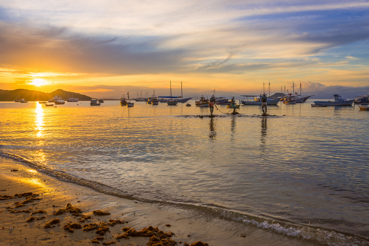 a fotografia mostra a praia de Manguinhos durante um pôr do sol em Rio de Janeiro Búzios. Diversos barcos estão ancorados na orla. O mar calmo e o céu em crepúsculo proporcionam uma bela paisagem.