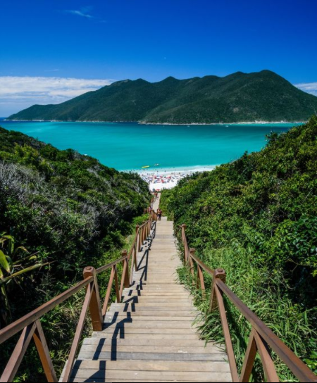 Descrição da imagem: a imagem mostra uma escadinha de madeira que leva a uma das praias de Arraial do Cabo. O final do caminho apresenta uma praia lindíssima com um mar azul turquesa.